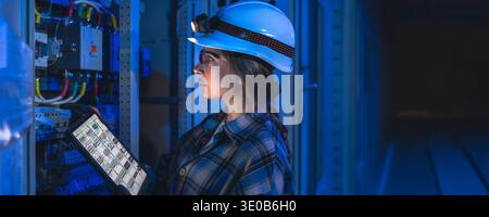 Female engineer inspecting electrical panel with tablet in dark industrial space. Engineer control. Stock Photo