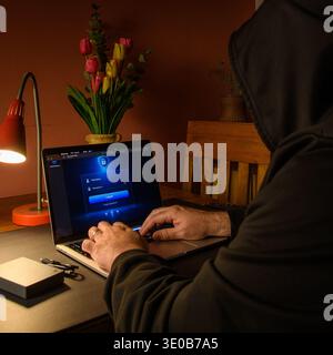 Hooded person typing on a laptop with login screen in a home office, conceptual image of cybersecurity, unauthorized access, hacking risk and online t Stock Photo
