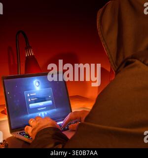 Hooded figure using a laptop with login screen under red light, conceptual image of cybercrime, hacking, suspicious login attempt and digital security Stock Photo