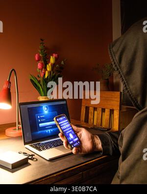 Hooded person holding a smartphone near a laptop login screen, conceptual image of two factor authentication, mobile verification and cybersecurity ac Stock Photo