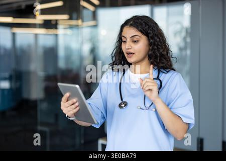 Young female medical professional in scrubs conducting a virtual patient consultation or medical training session using a tablet ,and gesturing during a video call in a modern office Stock Photo