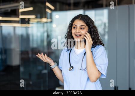 Young female healthcare professional wearing blue scrubs and a stethoscope, smiling and gesturing while talking on a smartphone, communicating in a modern medical setting Stock Photo