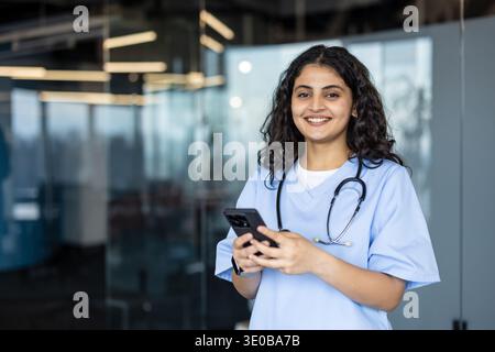 Young asian woman wearing blue scrubs and a stethoscope smiling, holding a smartphone while standing in a contemporary healthcare environment, representing communication and digital health Stock Photo