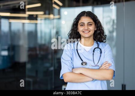Young female medical professional in scrubs and stethoscope smiling, standing with arms crossed at a modern clinic or hospital, representing healthcare, wellness, and dedicated service Stock Photo