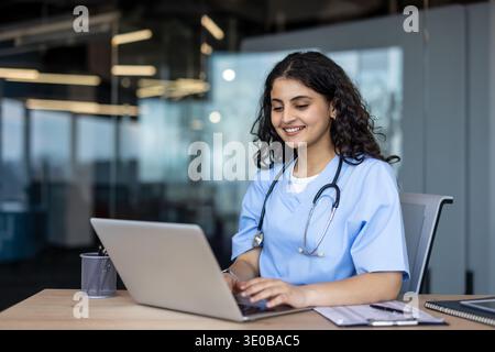 Young smiling female medical professional in scrubs and stethoscope working on a laptop at a modern clinic desk, typing and providing telehealth support and patient care remotely Stock Photo