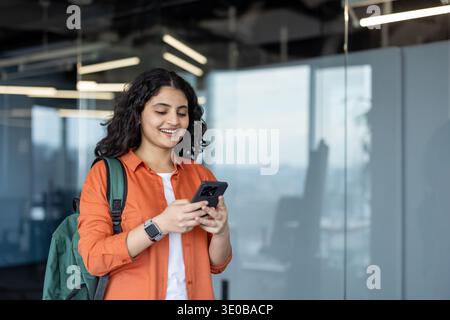 Young indian woman carrying a backpack and using a mobile phone, smiling while standing in a contemporary office or university hallway, engaging with digital communication and technology Stock Photo