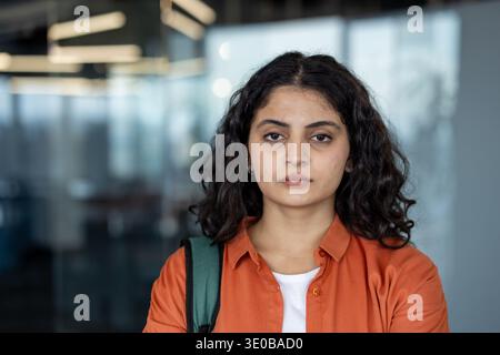 Young woman with curly hair and an orange shirt standing in a modern office, portraying professionalism, focus, and determination in a corporate environment Stock Photo