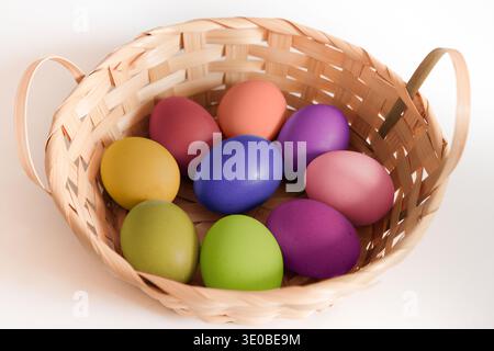 Colorful Easter eggs in a woven basket on a white background, featuring pastel shades of yellow, pink, purple, green, and blue. Stock Photo