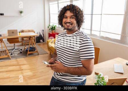 Young Indian man holding smartphone and smiling at camera at home office Stock Photo