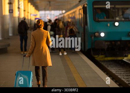 A woman in a tan coat walks toward a blue train on a station platform, pulling a light blue suitcase as she prepares to board for her next destination Stock Photo