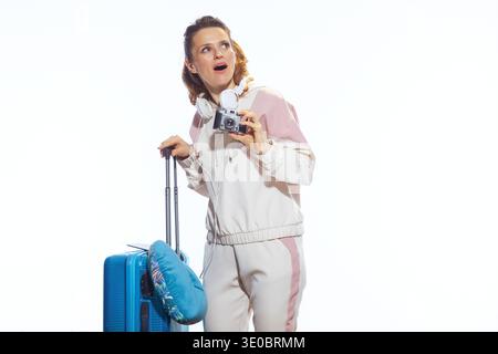 An excited woman in pink lounge wear looks up in surprise while holding a vintage camera and pulling her blue suitcase with a patterned travel pillow. Stock Photo