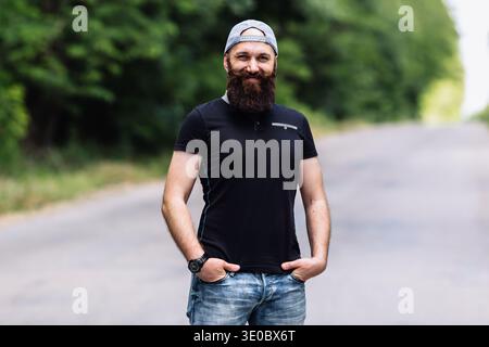 A handsome bearded man posing confidently on an empty road with his hands in pockets. He is wearing a grey baseball cap, black t-shirt, and jeans. Blu Stock Photo