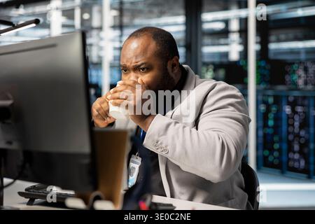 Data center admin drinking coffee and using PC to update servers tech, configuring gear. Man enjoying beverage in server farm, reviewing hardware analytics, ensuring reliable network operations Stock Photo