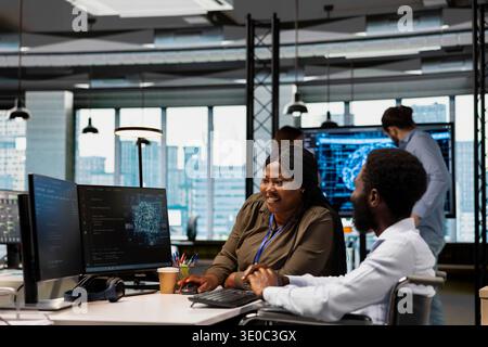 Smiling technician in wheelchair and coworker using AI automation, refining algorithms. Teamworking african american woman and system administrator with paraplegia using artificial intelligence Stock Photo