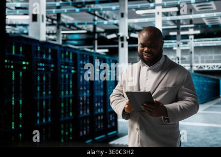 Smiling data center engineer using tablet to protect servers from exploitable software vulnerabilities, auditing systems. Happy cybersecurity expert using device, looking for bugs slowing down servers Stock Photo