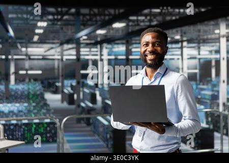 Portrait of cheerful black technician running diagnostics on AI systems, using machine learning equipment to identify errors or bugs. Confident man working to prevent failure and downtime. Stock Photo