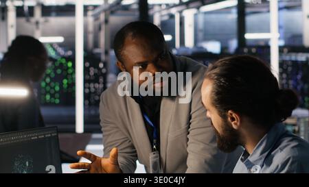 Server farm manager overseeing engineer examining neural network algorithms. Data center team leader and worker reviewing performance dashboards of artificial intelligence infrastructure, camera A Stock Photo