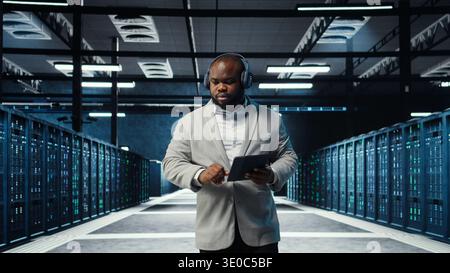 Server room programmer wearing headphones troubleshooting errors using software on tablet. IT specialist listening music, walking in data center fixing bugs using device , camera A Stock Photo