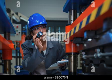 Indian female industrial engineer checks machinery with a flashlight while performing quality control in a manufacturing environment. Stock Photo