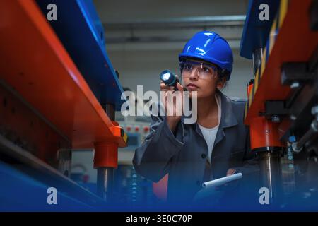Indian female industrial engineer uses flashlight to inspect machinery in a manufacturing factory for quality control purposes. Stock Photo