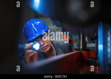 Indian female industrial engineer checks machinery using a flashlight in a manufacturing plant during quality control. Stock Photo
