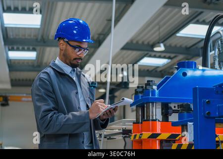 An African American industrial engineer inspects machines for quality control in a manufacturing plant while taking notes. Stock Photo