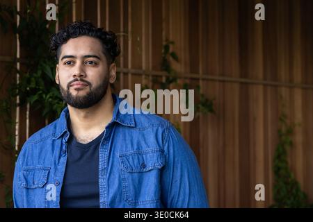 Man standing on patio against slatted wall with climbing vines, wearing denim shirt over black tee Stock Photo