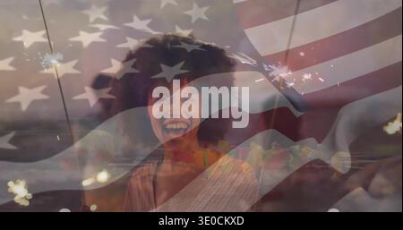 Laughing woman wearing light top holding sparkler, cheering at shore at dusk, American flag overlay Stock Photo