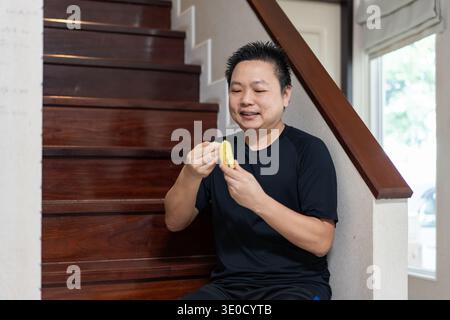 A man sits on the stairs at home, smiling while eating a banana as a healthy snack and source of natural energy after exercise. Stock Photo