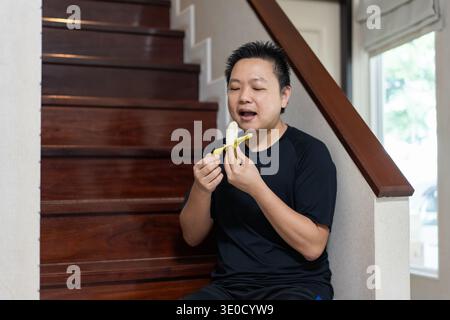 A man sits on the stairs at home, smiling while eating a banana as a healthy snack and source of natural energy after exercise. Stock Photo