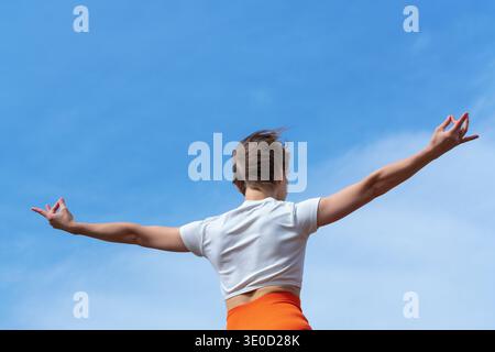 Back view of a woman with short hair practicing yoga outdoors. Her arms are spread wide in a gesture of freedom and meditation against a clear blue sk Stock Photo