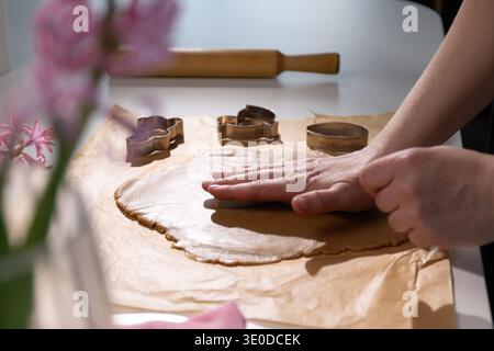 Hands press gingerbread cookie dough on baking paper, preparing Easter bunny and other shapes. Metal cookie cutters and a wooden rolling pin sit on a clean kitchen counter. Stock Photo