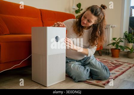 Woman sitting on the floor adjusting the display settings on a smart air purifier, bringing fresh air and better breathing to her modern home environment, prioritizing health technology and comfort Stock Photo