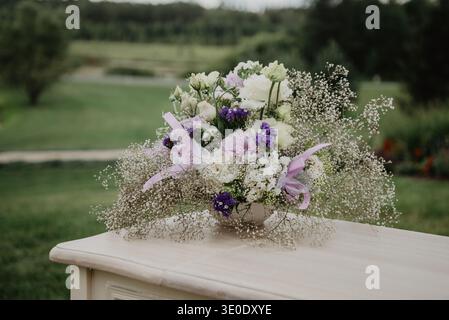 Delicate wedding flower arrangement with white and purple blossoms placed on an outdoor table. Soft natural light and blurred garden landscape create Stock Photo