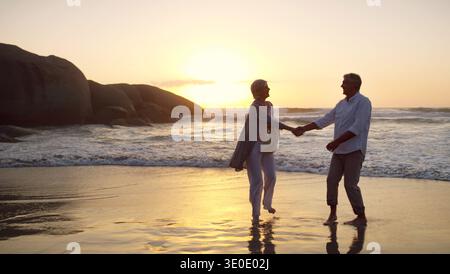 Elderly, couple and holding hands with sunset on beach for romantic holiday or weekend together. Senior, man and woman dancing with sunshine on ocean Stock Photo