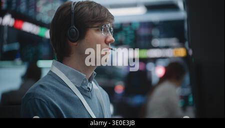 Close Up of Determined Young Specialist Deeply Engaged in Technical Analysis. Equipped With Professional Headset, Man Meticulously Monitors Fluctuating Charts, Global Indices Across Multiple Screens. Stock Photo