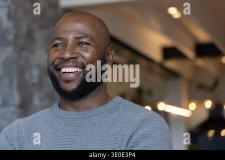 African American man smiling, wearing gray knit crewneck, standing near stone column in modern cafe Stock Photo