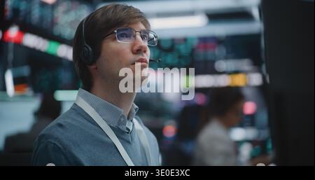 Close Up of Determined Young Specialist Deeply Engaged in Technical Analysis. Equipped With Professional Headset, Man Meticulously Monitors Fluctuating Charts, Global Indices Across Multiple Screens. Stock Photo