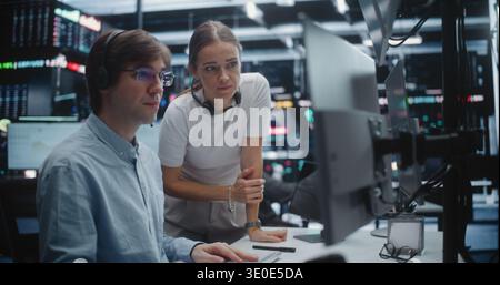 Female Manager Advising Young Male Trader in Busy Financial Office. Analyst With Supervisor Review Complex Analytics on Display, Gesturing Towards Key Data Points. Mentorship and Office Coordination. Stock Photo