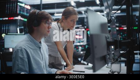 Female Manager Advising Young Male Trader in Busy Financial Office. Analyst With Supervisor Review Complex Analytics on Display, Gesturing Towards Key Data Points. Mentorship and Office Coordination. Stock Photo