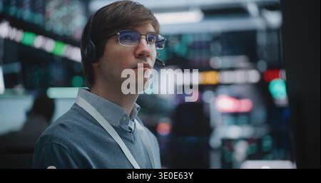 Close Up of Determined Young Specialist Deeply Engaged in Technical Analysis. Equipped With Professional Headset, Man Meticulously Monitors Fluctuating Charts, Global Indices Across Multiple Screens. Stock Photo