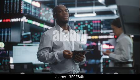 Senior Broker Analyzing Market Trends With Calm Confidence. Black Male Wearing Headset and Lanyard, Standing in High Tech Exchange, Holds Digital Tablet and Gazes Thoughtfully Toward Data Screens. Stock Photo