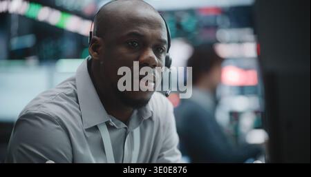 Close Up of Determined Young Specialist Deeply Engaged in Technical Analysis. Equipped With Professional Headset, Man Meticulously Monitors Fluctuating Charts, Global Indices Across Multiple Screens. Stock Photo