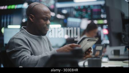 African American Investor Tracking Asset Performance in Innovative Trading Center. Man Focused on Digital Dashboard on Monitors and Digital Tablet. Intense Concentration in Global Finance Industry. Stock Photo