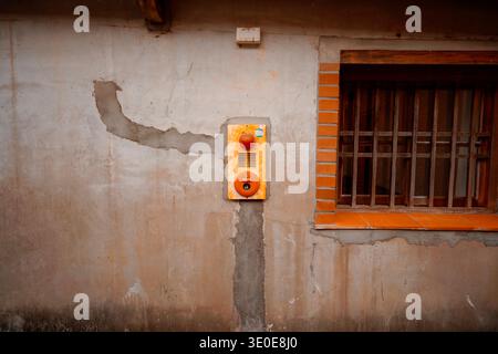 Rusty alarm bell mounted on weathered gray wall of traditional Taiwanese architecture, with cracked plaster, brick window frame, and wooden bars. Stock Photo