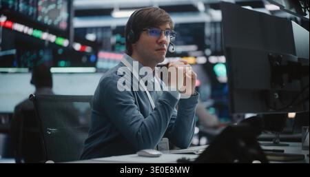 Young Male Financial Analyst in Glasses and Headset Sits at Modern Workstation, Gazes at Large Trading Monitors, Tracking Real Time Price Movements During Critical Market Session. Digital Monitoring. Stock Photo