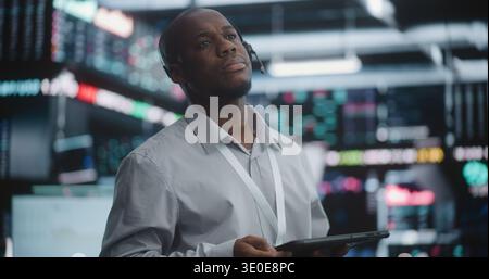 Senior Broker Analyzing Market Trends With Calm Confidence. Black Male Wearing Headset and Lanyard, Standing in High Tech Exchange, Holds Digital Tablet and Gazes Thoughtfully Toward Data Screens. Stock Photo