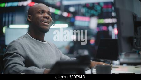 Serious African American Financial Professional Monitoring Stock Market. Male Trader at High Tech Workstation, Intently Watching Real Time Stock Charts While Reporting on Market Changes Via Headset. Stock Photo