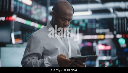Senior Broker Analyzing Market Trends With Calm Confidence. Black Male Wearing Headset and Lanyard, Standing in High Tech Exchange, Holds Digital Tablet and Gazes Thoughtfully Toward Data Screens. Stock Photo
