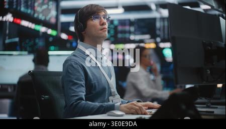 Young Male Financial Analyst in Glasses and Headset Sits at Modern Workstation, Gazes at Large Trading Monitors, Tracking Real Time Price Movements During Critical Market Session. Digital Monitoring. Stock Photo